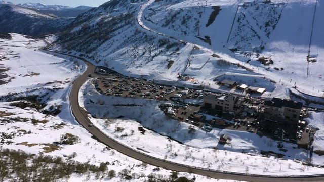 Aerial, tracking, drone shot of the Leitariegos ski resort, a parking lot full of cars, hotels and people skiing on the mountains, on a sunny day, in Leon, Spain