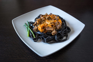 black pasta with cuttlefish ink. Pasta of durum wheat semolina with squid ink with beef and sweet pepper sauce on a square plate decorated with green onions with fork