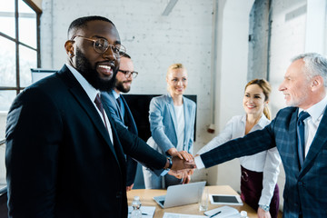 cheerful african american man looking at camera while putting hands together with coworkers