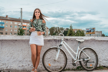 Happy girl is standing in the summer in city in hand a phone and a cup of coffee. Near parked bike. Lifestyle in the city. Emotions of joy fun and rest after work. Free space for text.