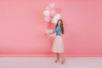 Full-length portrait of attractive stylish girl with gift wearing high heel shoes and midi skirt for friend's birthday party. Adorable curly young woman holding present box with balloons for event