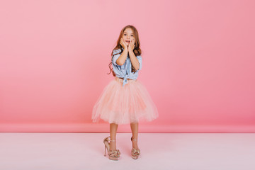 Charming cute little girl with long brunette hair in tulle skirt and blue shirt expressing isolated on pink background. Having fun in mothers shoes of funny joyful child, cheerful mood