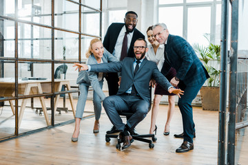 cheerful businessman sitting on chair with outstretched hands near multicultural coworkers