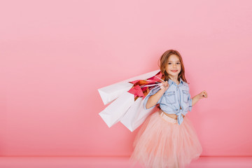 Amazing cute little girl with long brunette hair walking with white packages isolated on pink...
