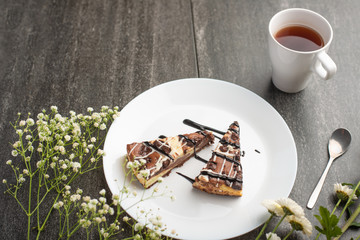On the plate, two slices of cheesecake with tea and flowers, for congratulations on a dark background