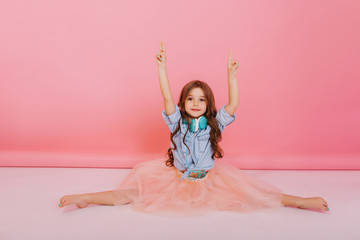 Sweet moments happy childhood of amazing young girl in tulle skirt making qymnastics split on white floor on pink background. Cute fashionable child with long brunette hair, blue headhones on neck