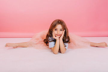 Leisure time of joyful charming little girl making gymnastics split on floor on pink background....