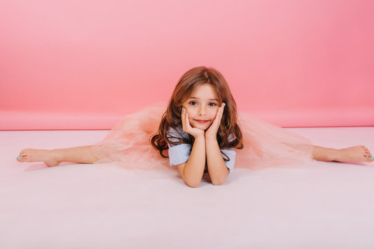Leisure time of joyful charming little girl making gymnastics split on floor on pink background. Elastic cute child in tulle skirt  with long brunette hair smiling to camera, expressing cheerful mood