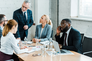 selective focus of african american man talking on smartphone near laptop and coworkers