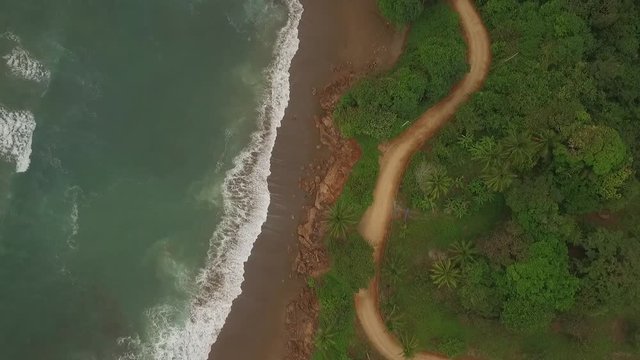 Aerial view of interesting dirt road between ocean waves and central american jungle moves bottom to top of scene