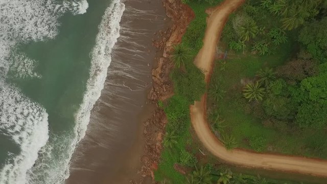 Aerial view of interesting dirt road between ocean waves and central american jungle with camera raise