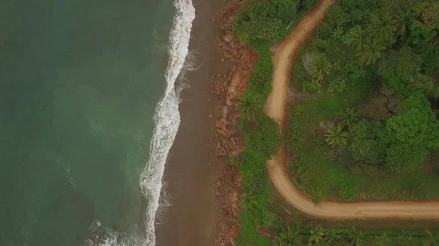 Aerial view of interesting dirt road between ocean waves and central american jungle with camera drop