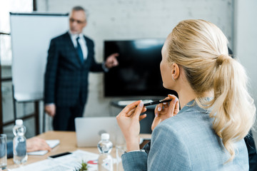 selective focus of blonde businesswoman holding pen while looking at business coach