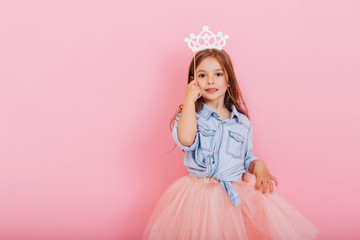 Pretty sweet little girl with long brunette hair in tulle skirt holding white crown on head isolated on pink background. Beautiful joyful child expressing true emotions. Place for text © Look!