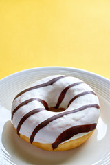 One single round donut with white ice glazing and stripes of dark brown chocolate on a white plate against a yellow background with copy space. Vertical image