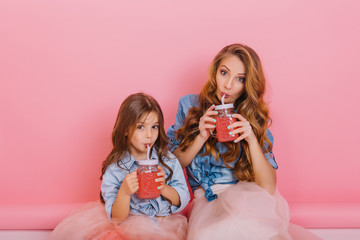 Mom and daughter in vintage clothes prepared tasty berry fresh and drink it from big glass jars sitting on the floor. Little curly girl posing with her adorable mother holding glass of cherry cocktail