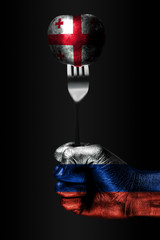 A hand with a drawn Russia flag holds a fork, on which is a ball with a drawn Georgia flag, a sign...