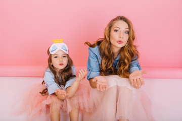 Graceful young woman in denim shirt and lush skirt having fun with her cute daughter in sleep mask at party. Little curly girl in stylish attire funny posing with elder sister on pink background