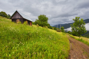 Summer mountain panorama. Small wooden house cottage and barn on green mountain valley on bright foggy sky, clouds and mountain