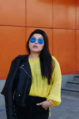 Portrait of a young brunette plus size woman. Audacity pan-Asian girl in a biker leather jacket with sunglasses poses near a bright wall. Body positivity movement. Street fashion. Leisure time