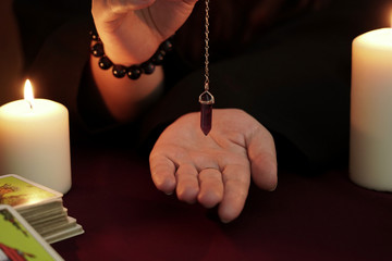 Witch is fortune teller in black robe holds amethyst pendulum on chain above hand. Tarot cards, amethyst stone, white candles on dark mystic background. Occult, esoteric, divination and wicca concept.