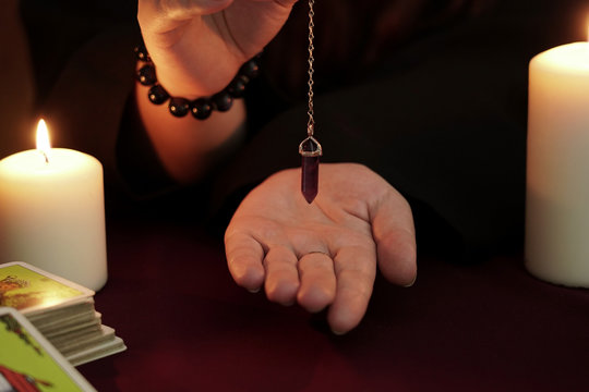 Witch is fortune teller in black robe holds amethyst pendulum on chain above hand. Tarot cards, amethyst stone, white candles on dark mystic background. Occult, esoteric, divination and wicca concept.