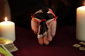 Witch in a robe holds on palm black and white dolls tied with red thread. Black on top. Tarot cards, ancient runes and white candles. Occult, esoteric, divination and wicca concept. Mystic background.