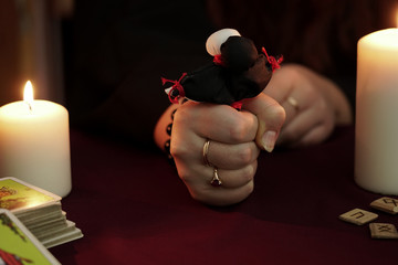 Witch in robe with rings clenches in her fist black and white dolls tied with red thread. Tarot cards, ancient runes and white candles. Occult, esoteric, divination and wicca concept. Dark background.