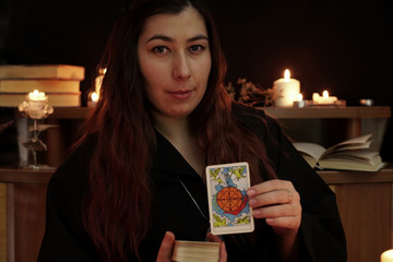 A witch is fortune teller with brown hair in black mantle shows a card. Magical ritual. Tarot cards, white candles on table. Occult, esoteric, divination and wicca concept. Mystic background.
