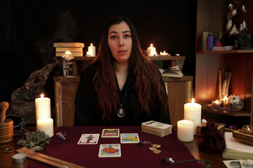 Portrait of witch in a black robe. Magical ritual. Tarot cards, white candles, aroma stick, ancient book and runes on table. Occult, esoteric, divination concept. Mystic background.