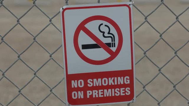 A quick zoom in on a red and white no smoking sign that is behind a chain linked fence.
