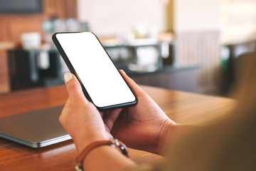 Mockup image of woman's hands holding black mobile phone with blank white screen with laptop computer on wooden table