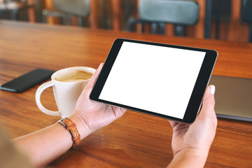 Mockup image of hands holding black tablet pc with blank white screen with coffee cup on wooden table