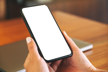 Mockup image of woman's hands holding black mobile phone with blank white screen with laptop computer on wooden table