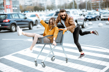 Young stylish coupe having fun riding with shopping cart on the outdoor parking near the supermarket