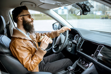 Stylish bearded man having a snack with tasty sandwich while driving a car in the city