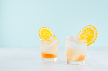 Oranges cold alcohol drinks with liquor layers, sliced orange, ice cubes in misted shot glass on pastel blue background.