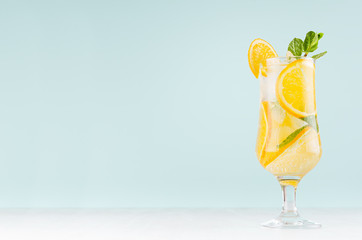 Oranges detox water with sliced orange, ice cubes, green mint in misted wineglass on pastel blue background, white wood board.