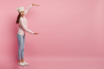 Indoor shot of happy Caucasian lady measures something big with two hands, dressed in white headgear, sweatshirt and jeans, isolated over pink background with empty space for your advertisement