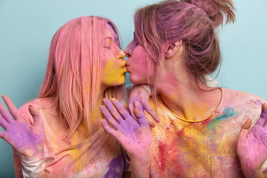 Studio shot of pleased two European young women kiss each other, smeared with colorful Holi powder, celebrate Indian spring holiday together, show colorful palms, isolated on blue background.