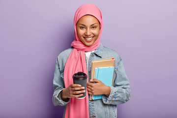 Cheerful Muslim woman in pink hijab, denim coat, carries pocketbook, spiral diary and takeaway coffee, has break after lecture isolated over purple background. Students life, religion and ethnicity