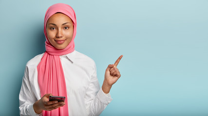 Beautiful calm dark skinned Muslim girl stands with mobile phone, wears pink veil and white shirt, points aside with index finger, shows direction to electronics shop. People, religion, technology