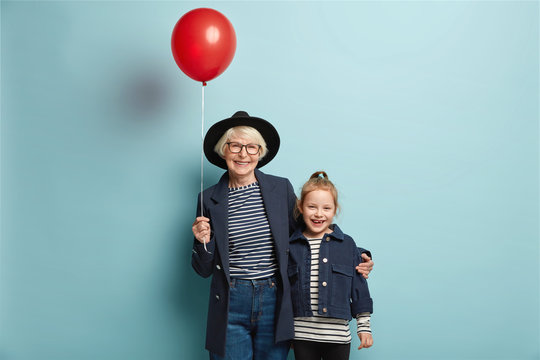 Studio shot of cheerful granddaughter and grandmother embrace together, come on party, hold red balloon, wear fashionable attire, have positive smiles on faces. Two generations. Relationships