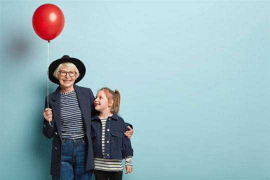 Indoor shot of fashionable senior woman embraces little child, enjoy spending time together, celebrate first day at school, hold air balloon, pose over blue background with blank space on right