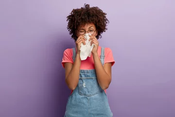 Fotobehang Portrait of sick African American woman sneezes in white tissue, suffers from rhinitis and running nose, has allergy on something, looks unhealthy, feels unwell. Symptoms of cold or allergy. © Wayhome Studio