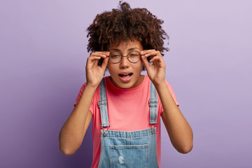 Young Afro girl keeps hands on frame of glasses, tries to concentrate, has bad vision, looks into distance, reads text with small letters opens mouth from interest focused at camera looks scrupulously