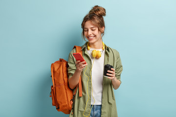 Photo of positive young woman in casual clothes, downloads multimedia file on cell phone, has headphones on neck, holds disposable cup with cappuccino, carries backpack, isolated on blue wall - obrazy, fototapety, plakaty