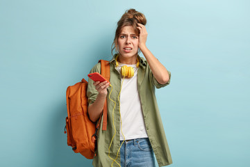 Isolated shot of dissatisfied schoolgirl holds cell phone connected to headphones, has frustrated expression, can not listen music without internet. Oh no, my songs disappeared from playlist