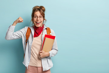 Obraz premium Optimistic happy college student with European appearance, raises clenched fists, shows muscles, celebrates successfully finished school year, holds books, has backpack, poses indoor with copy space