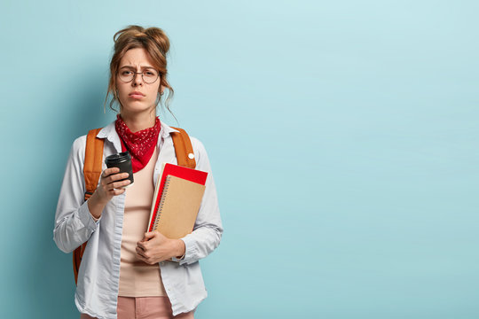 Dissatisfied sleepy student poses with books, notepad and coffee, feels overworked preparing for exam, has tired facial expression, wears optical glasses and casual clothes, caries rucksack on back